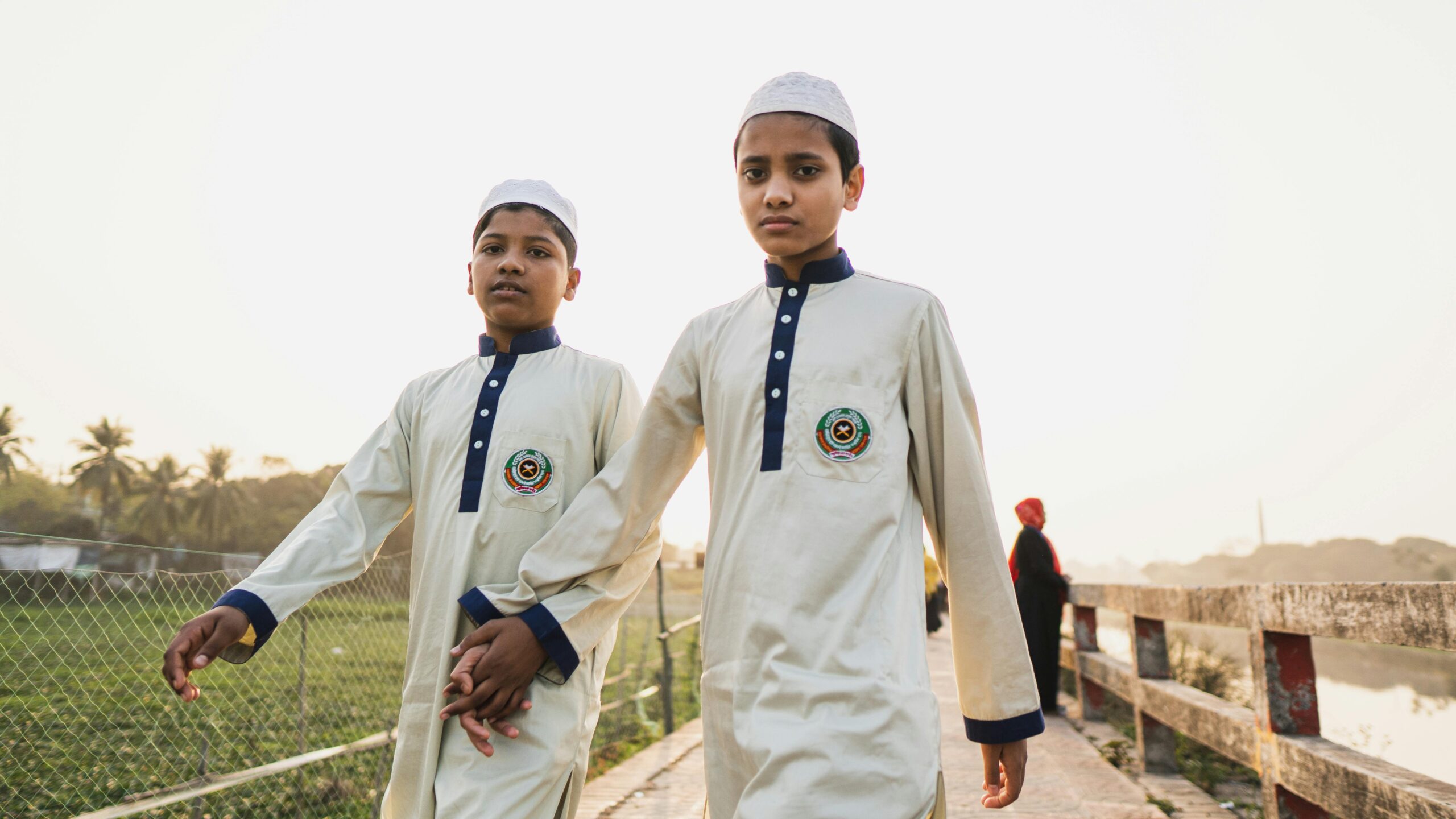 Two young boys in traditional Islamic school uniforms walking hand in hand on a sunny day.
